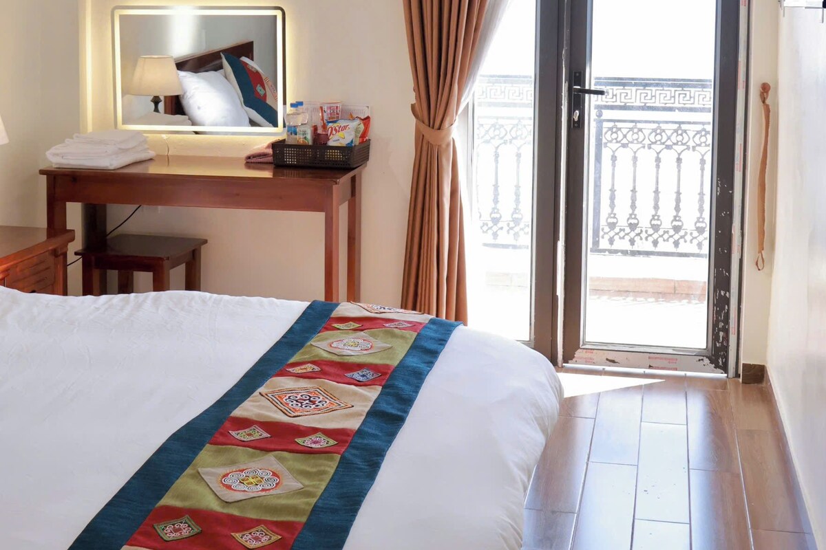 Habitación de hotel con cama, escritorio con espejo, ventana con cortinas y vista al balcón.