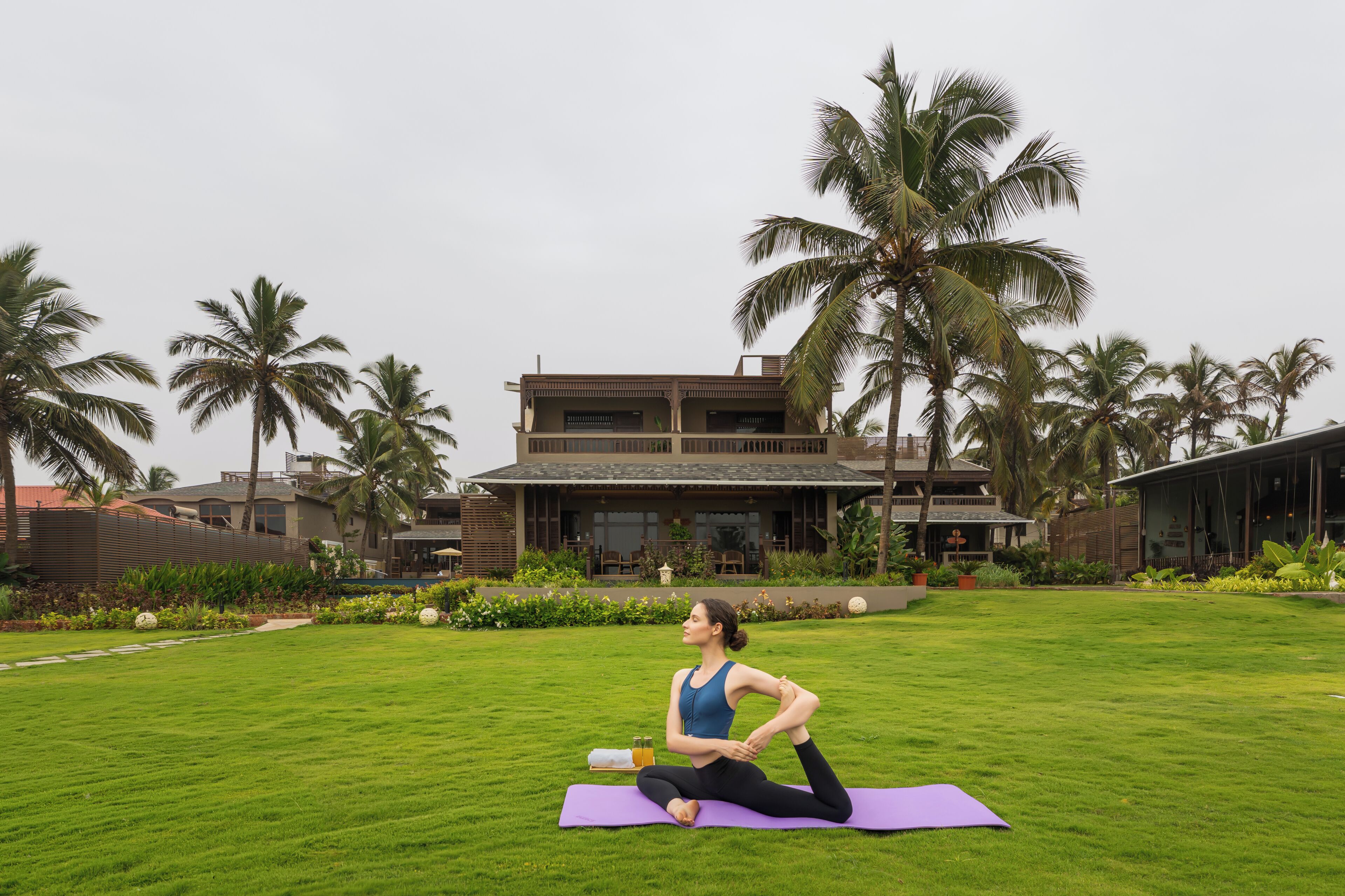 A person performing a yoga stretch on a purple mat in a grassy area with palm trees and a building in the background.