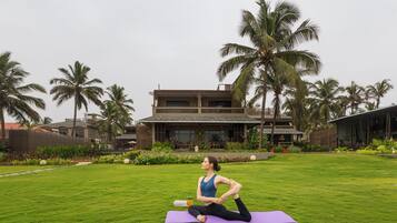 A person performing a yoga stretch on a purple mat in a grassy area with palm trees and a building in the background.