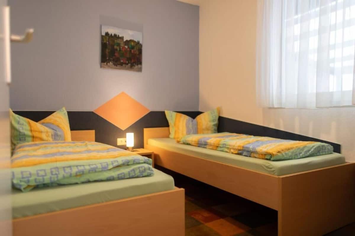 A room with two beds, a nightstand with a lamp, a wall-mounted picture, and a window with sheer curtains.