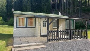 A small cabin with a porch, a fenced yard, and a forested backdrop.