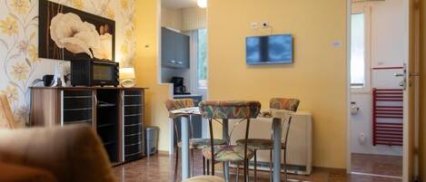 A living room with a sofa, a dining table with chairs, a TV, and a kitchen area.
