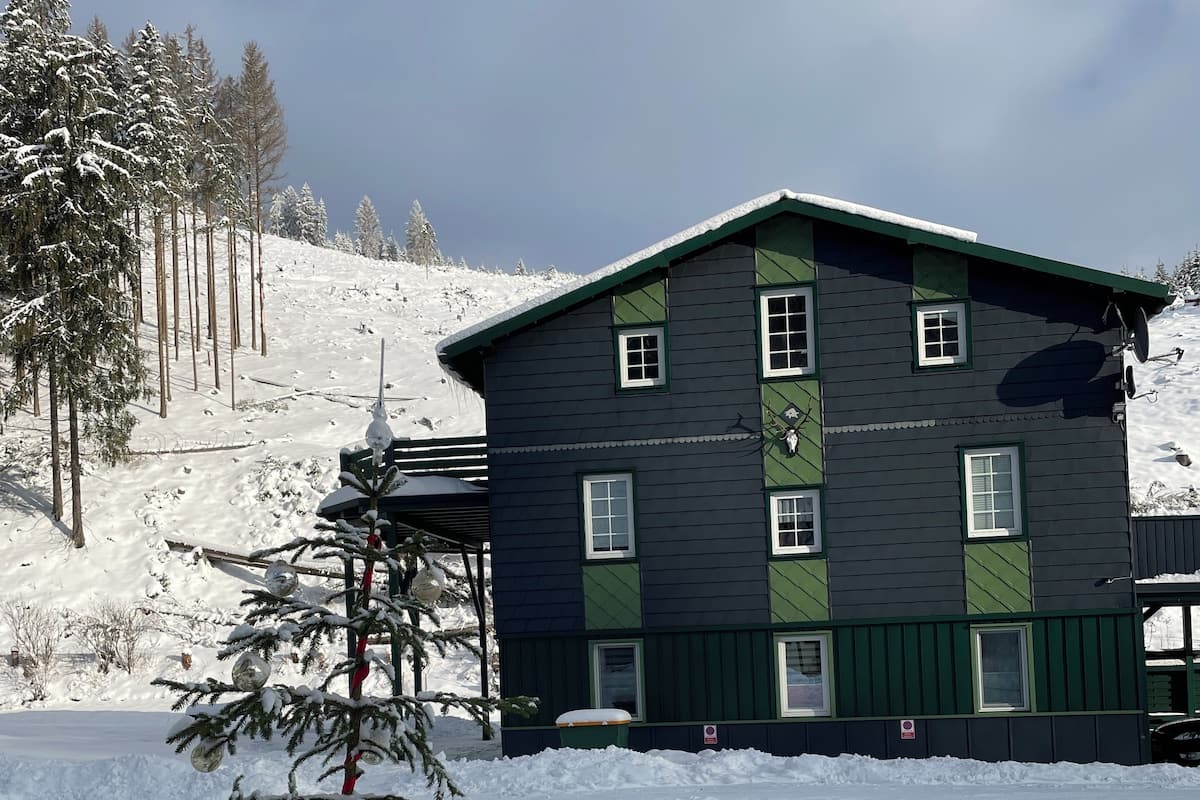 A two-story building with a dark exterior and green trim, surrounded by snow-covered ground and trees.