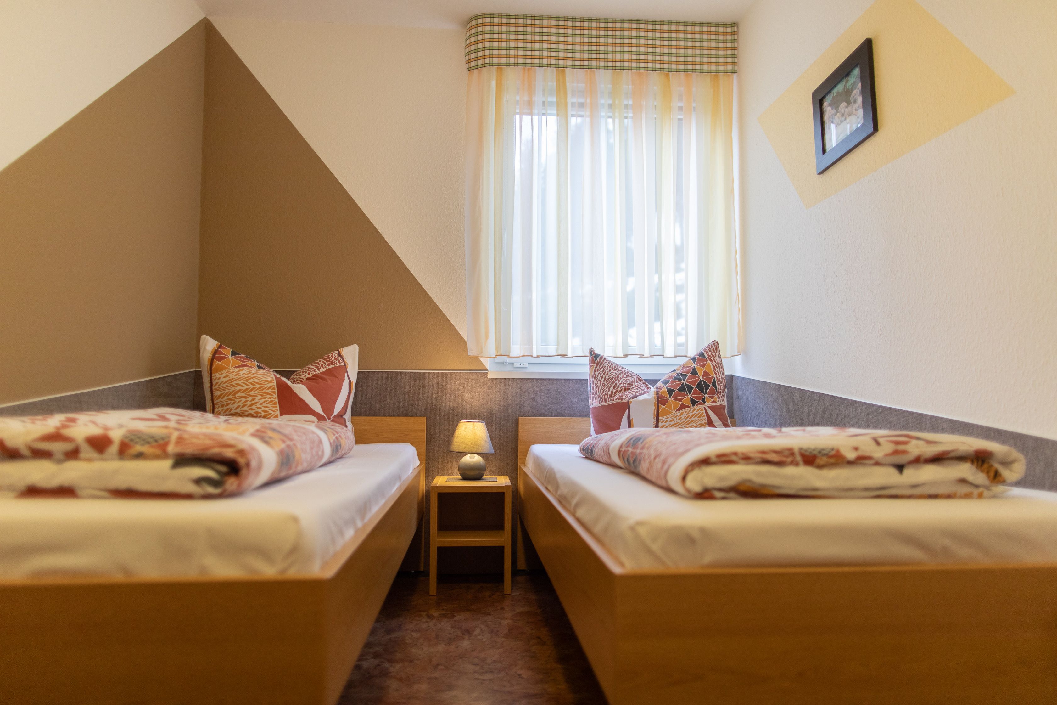A small, triangular room with two beds, a nightstand with a lamp, and a window with sheer curtains.
