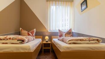 A small, triangular room with two beds, a nightstand with a lamp, and a window with sheer curtains.