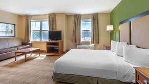 A hotel room with a bed, a television on a stand, a sofa, a small table, and a window with curtains. - Studio 6 Gaithersburg|1 Queen|Nmsk|Sofa-Bed|Kitchen (Baltimore MD)