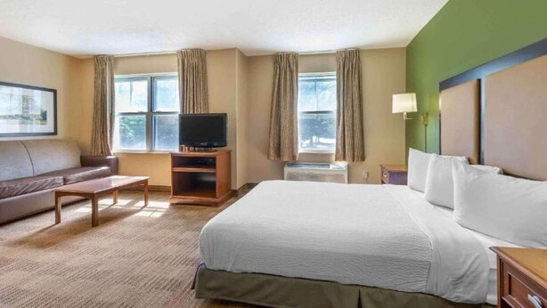 Una habitación de hotel con una cama, un televisor en un mueble bajo, un sofá, una mesita y una ventana con cortinas.