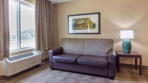 Habitación de hotel con sofá, un cuadro enmarcado en la pared, una lámpara en una mesita de noche y una ventana con cortinas.
