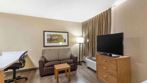 A hotel room with a sofa, a small wooden table, a TV on a wooden stand, a desk with a chair, a framed picture on the wall, and a lamp. - Studio6 Fishkill 2 Full Beds, Mobil Tub, Nsmk Kitchen (Fishkill)