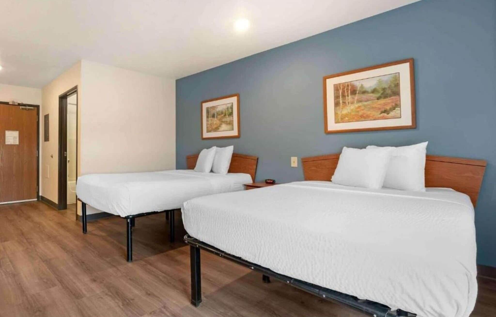 A hotel room with two beds, wooden flooring, and two framed landscape paintings on the walls.