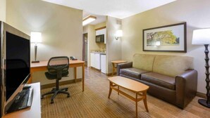 Une chambre d’hôtel comprenant un canapé, un bureau avec une chaise, une télévision, une petite table, un coin cuisine et un tableau encadré au mur.