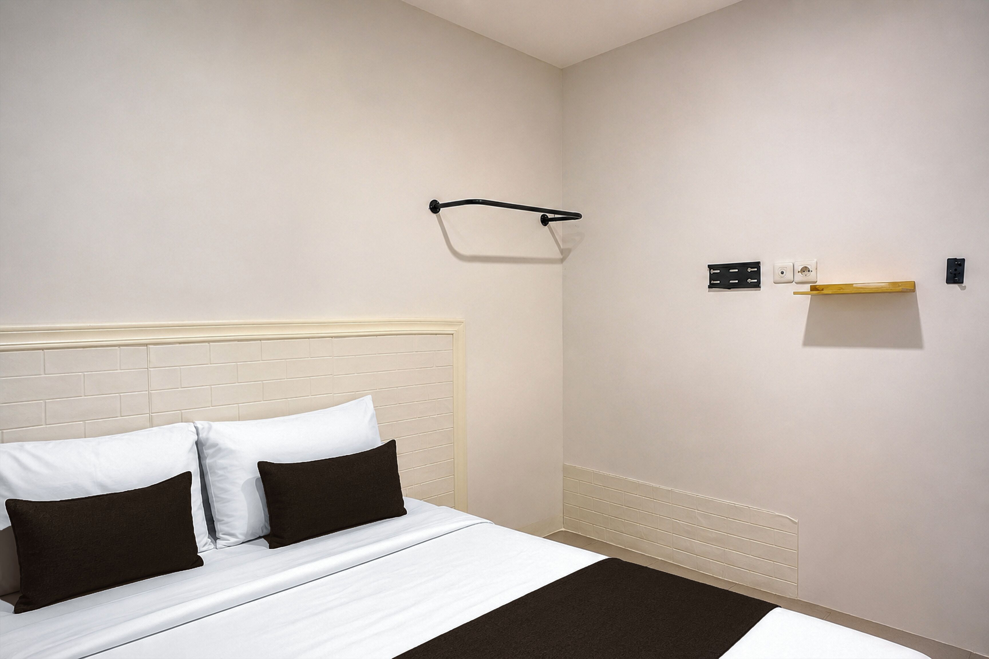 A hotel room with a bed, a headboard, a wall-mounted towel rack, and a small shelf with a light fixture.