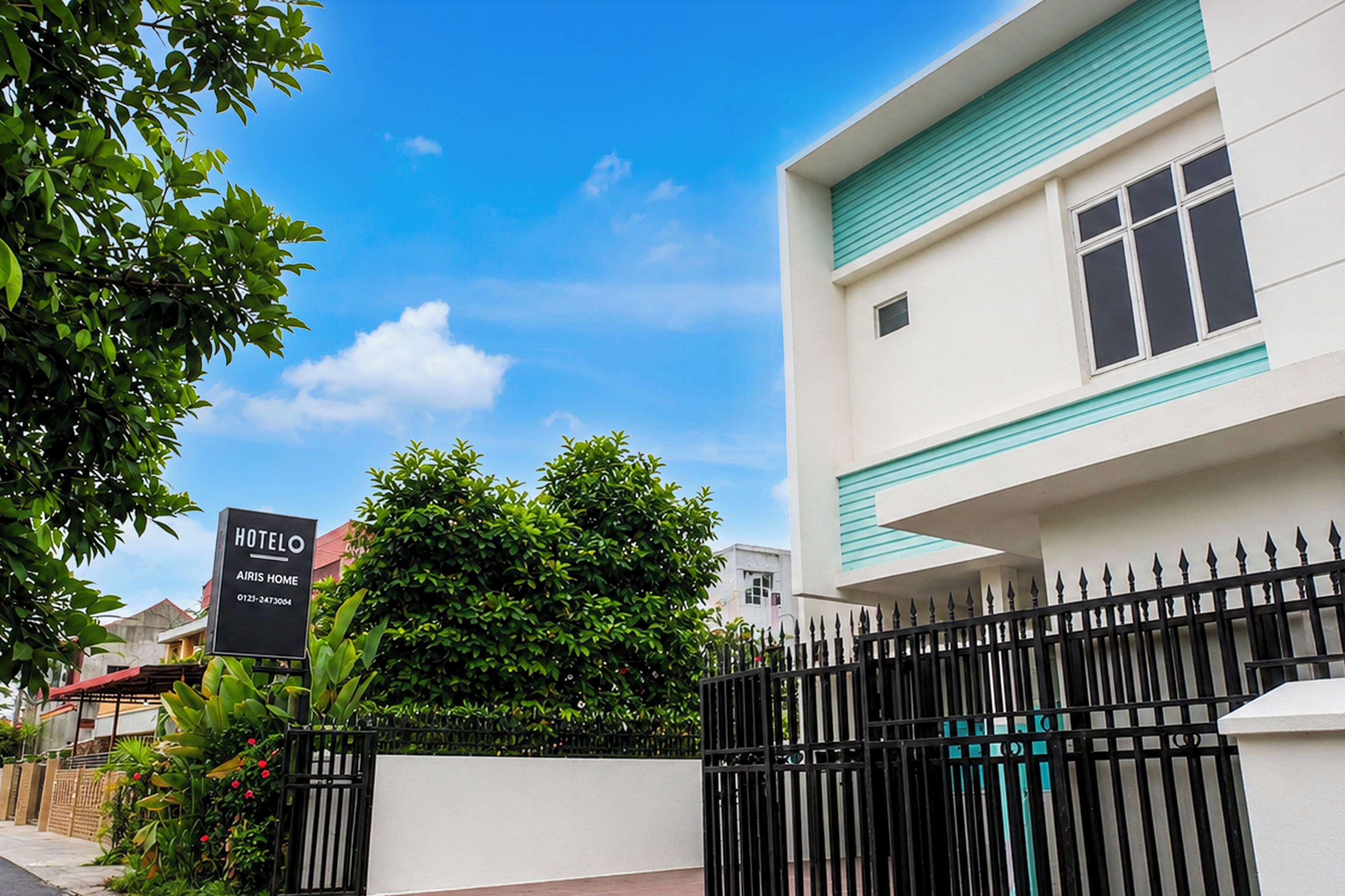 A hotel named 'HOTEL AJRIS HOME' with a signboard, a gated entrance, and a building with a white facade and teal trim.