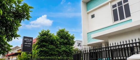 A hotel named 'HOTEL AJRIS HOME' with a signboard, a gated entrance, and a building with a white facade and teal trim.