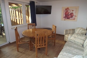 A dining area with a wooden table and chairs, a sofa, a TV on the wall, and a window with a view of a wooden fence.