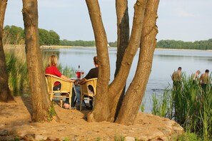 Ein Seeufer mit Tisch und Stühlen unter Bäumen, mit Blick auf einen stillen See.