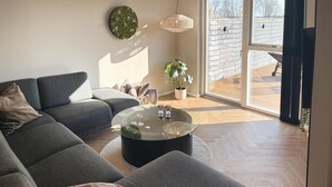 A modern living room with a sectional sofa, a glass coffee table, a hanging light fixture, and a large window offering a view of the outdoors. - Pet friendly home in Børkop with sauna (Børkop)