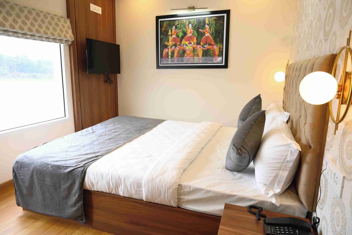 A hotel room with a bed, pillows, a bedside table with a phone, a wall-mounted TV, and a framed picture on the wall.