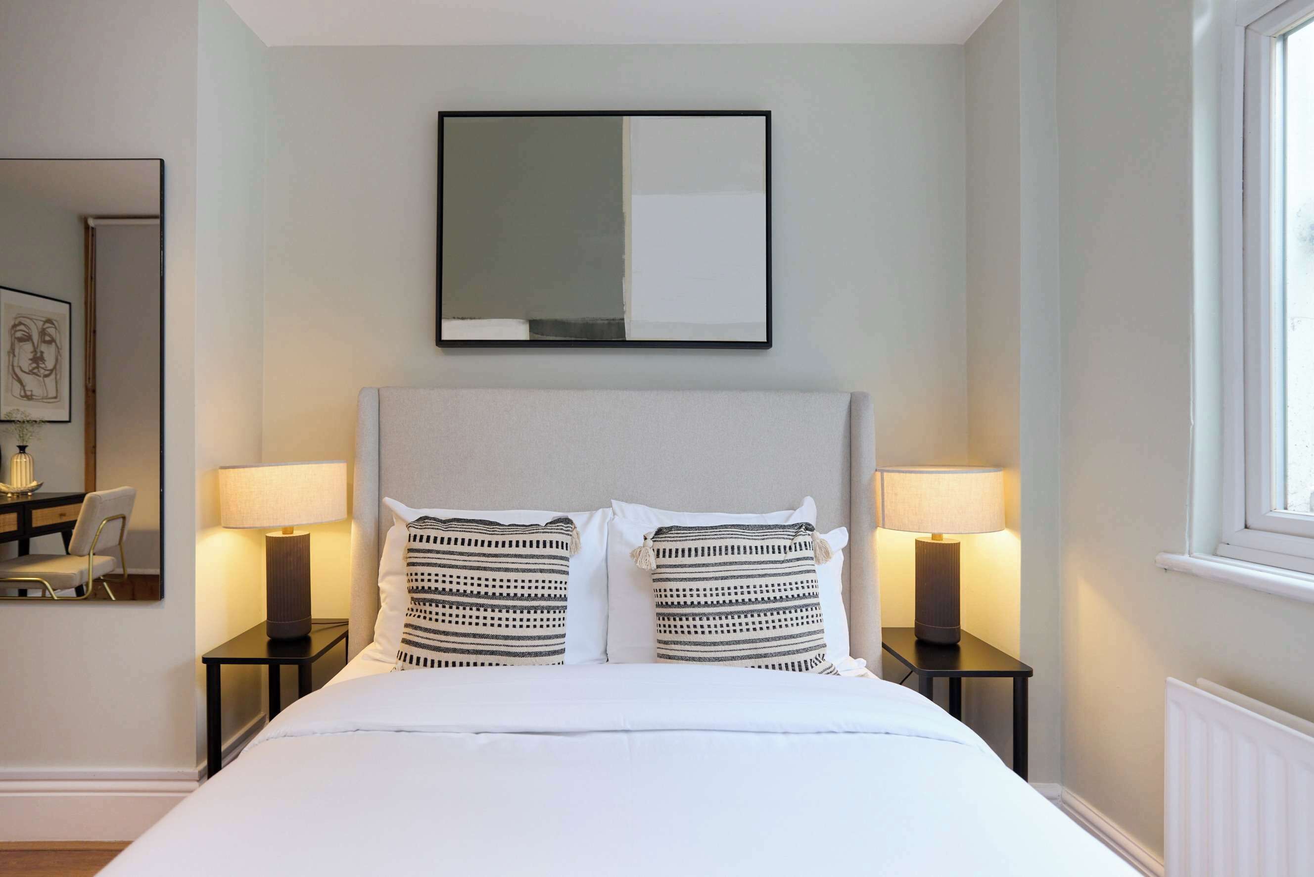 Habitación de hotel ordenada con cama, dos lámparas de noche, un espejo grande y una ventana.