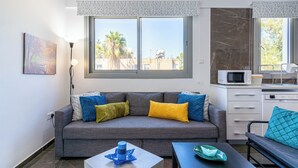 A modern living room with a grey sofa, colorful cushions, a coffee table with a blue object, a small table with a green object, and a kitchen area with a microwave. - Kmeidos 1-BR Apt & Balcony in Larnaca (Larnaca)