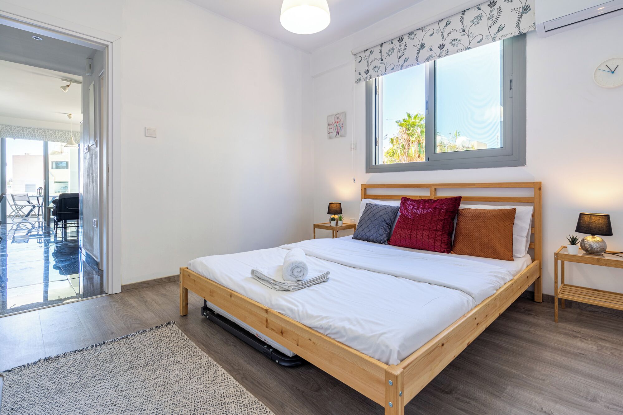 A bedroom with a wooden bed, a window with a view, a small bedside table, and a door leading to a balcony.