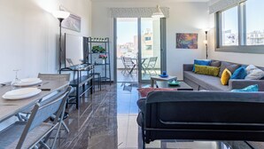 A modern living room with a sofa, a dining area with a table and chairs, a balcony with a view of buildings, and a TV mounted on the wall. - Kmeidos 1-BR Apt & Balcony in Larnaca (Larnaca)