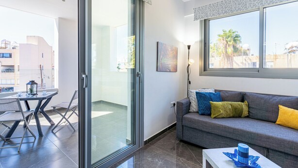 A modern living room with a grey sofa, a glass door leading to an outdoor area, a table with chairs, and a large window with a view of buildings and trees. - Kmeidos 1-BR Apt & Balcony in Larnaca (Larnaca)
