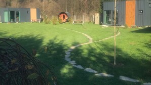 Een gazon met een klein gebouw met een rood dak, een houten bankje en een pad dat leidt naar een groter, grijs gebouw met een oranje streep.