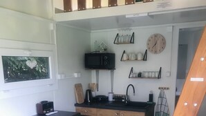 Een compacte keuken met een magnetron, spoelbak en een kleine koelkast. Er is een raam met een hartdecoratie en een klok aan de muur.