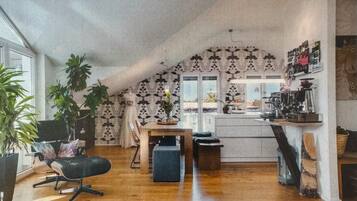Ein gemütliches Loft-Zimmer mit Holzboden, einem Esstisch, einem schwarzen Stuhl, einer kleinen Sitzecke und einer Küchenzeile mit Arbeitsplatte und verschiedenen Gegenständen.