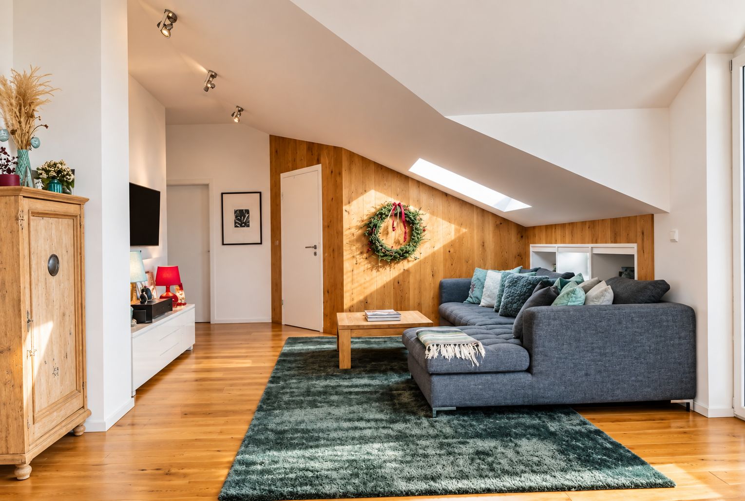 Ein modernes Wohnzimmer mit einem grauen Sofa, Holzboden, einem grünen Teppich, einem Holzschrank und einem an der Wand befestigten Fernseher.