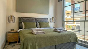 A modern bedroom with a large window, a bed with green bedding, two bedside lamps, and a small bedside table. - Qoople Smart 4 Cleopatra 7 Family 2+1 Apartment (Alanya)