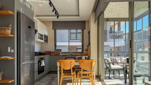 A modern kitchen with a built-in refrigerator, a dining area with two wooden chairs, and a view of the city through large windows. - Qoople Smart 4 Cleopatra 7 Family 2+1 Apartment (Alanya)