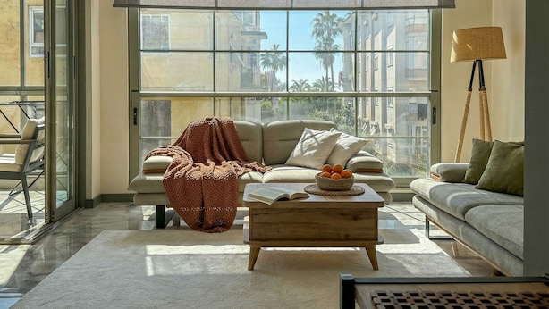 A spacious living room with a large window, a sofa, a coffee table, and a floor lamp. - Qoople Smart 4 Cleopatra 7 Family 2+1 Apartment (Alanya)