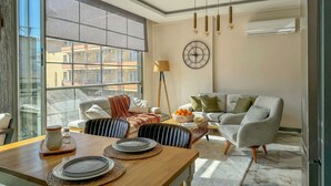 A modern living room with a dining area, a large window, a clock, and a sofa. - Qoople Smart 4 Cleopatra 7 Family 2+1 Apartment (Alanya)