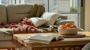 A cozy living room with a sofa, cushions, a knitted blanket, a wooden side table with a book and a basket of oranges. - Qoople Smart 4 Cleopatra 7 Family 2+1 Apartment (Alanya)