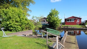A calm waterway with a dock, a green boat, and a red building in the background. - Möwenglück am Meer - Holiday home Möwenglück am Meer with garden by the water (Südbrookmerland)