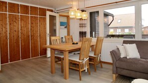 A dining area with a wooden table and chairs, a brown sofa, and a window with a view of houses. - Möwenglück am Meer - Holiday home Möwenglück am Meer with garden by the water (Südbrookmerland)
