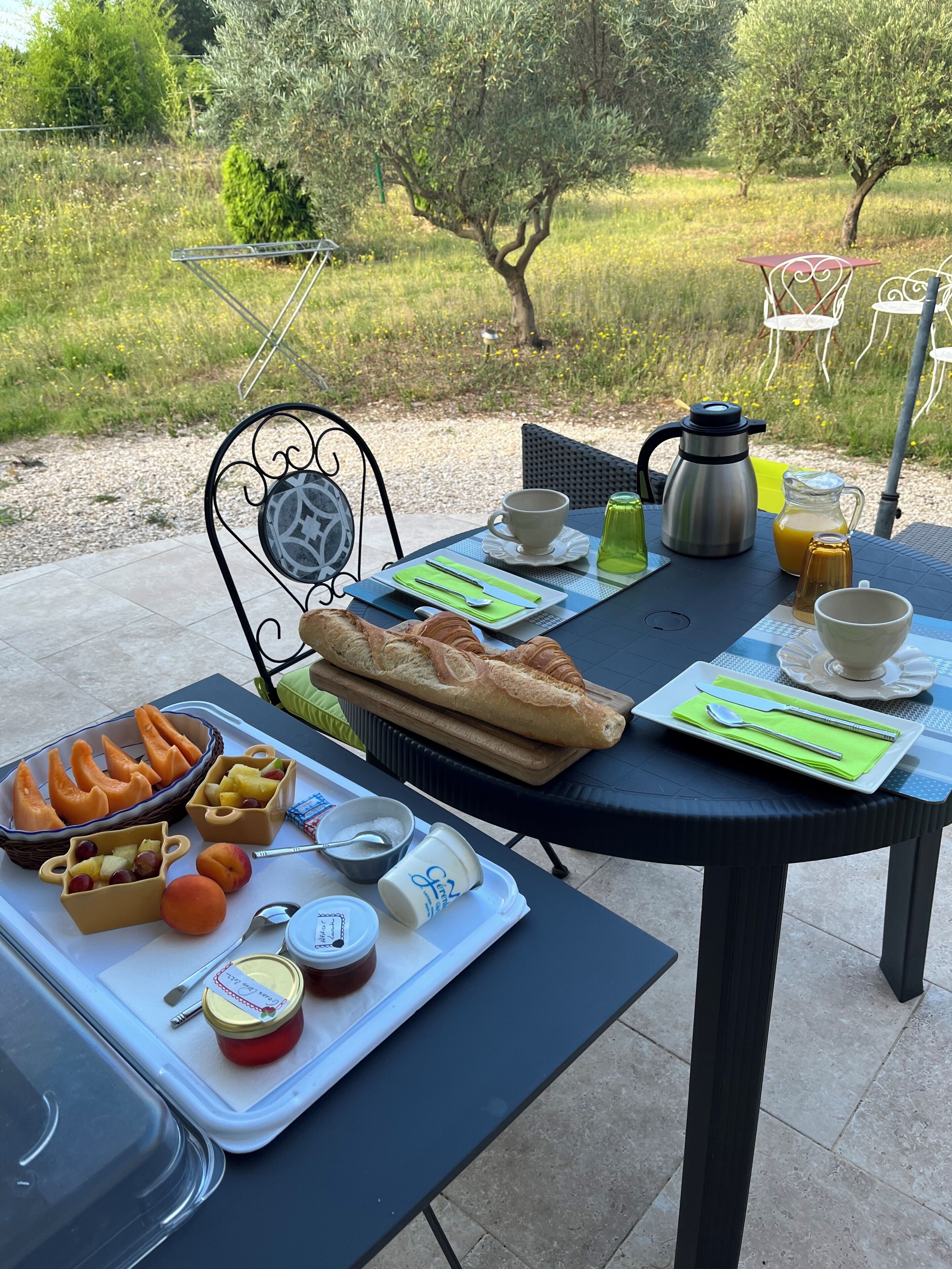 An outdoor dining table with a croissant, fruit, and condiments, set against a garden backdrop.
