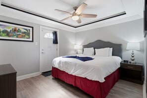 Ein Hotelzimmer mit einem Bett, einem Deckenventilator, einem Nachttisch mit Lampe, einer Tür und einem gerahmten Bild an der Wand.