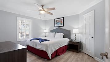 Ein Schlafzimmer mit Bett, Nachttischen, Deckenventilator, Fenster mit Jalousien und einer Tür.