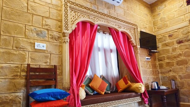 A room with a stone wall, a wooden bench with cushions, a television, and a table with a kettle. - Hotel Unique Heritage  (Jaisalmer)