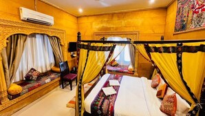 A room with a four-poster bed, a bench with cushions, a chair, and a ceiling fan. - Hotel Unique Heritage  (Jaisalmer)