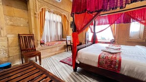 A room with a bed, a wooden chair, a table, and a red canopy bedspread. - Hotel Unique Heritage  (Jaisalmer)