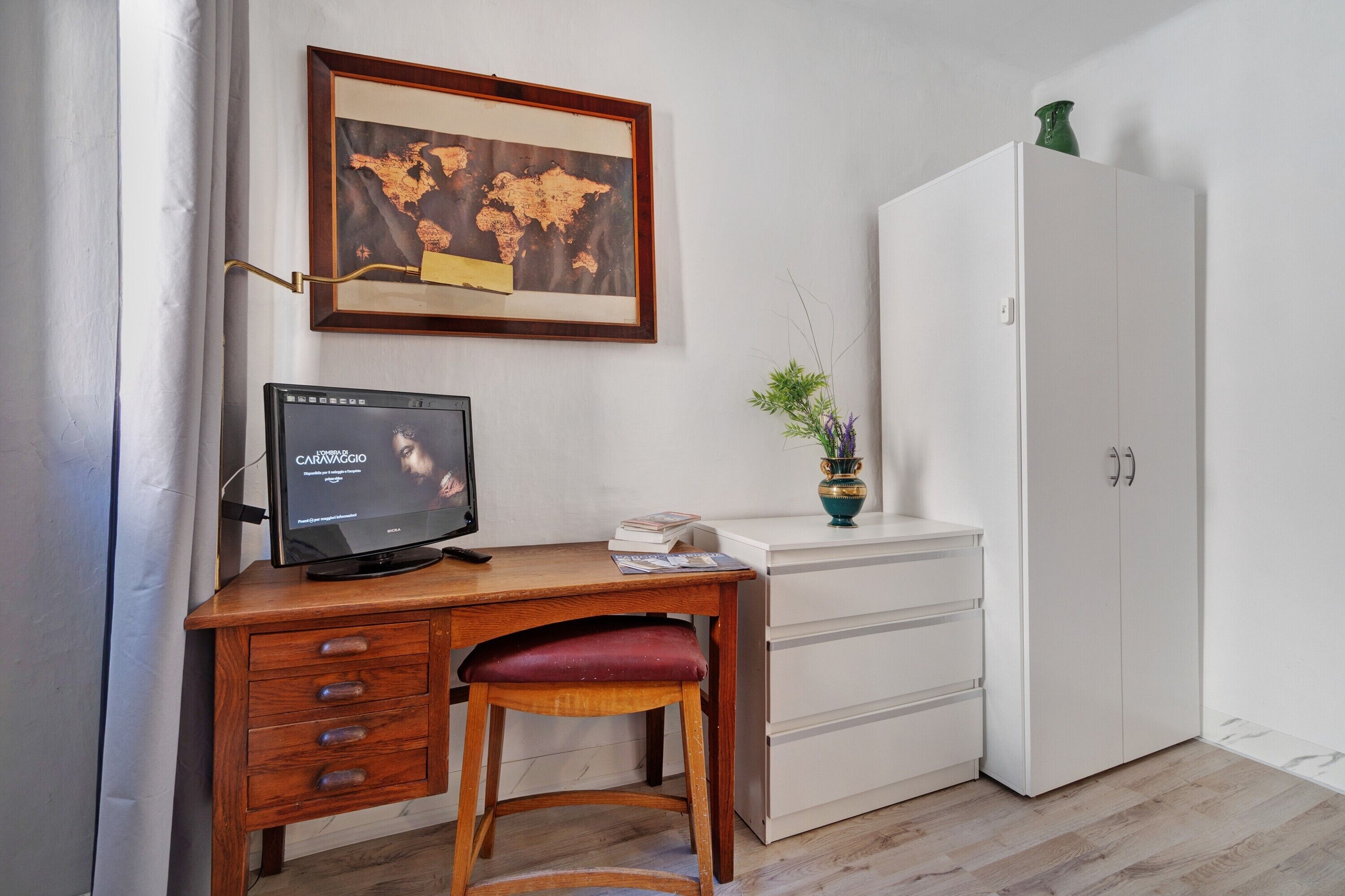 A room with a wooden desk, a television, a white wardrobe, and a framed map on the wall.