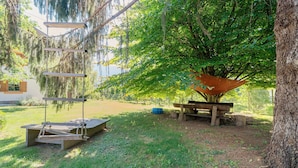 Ein hölzernes Schaukelgestell, ein Picknicktisch und eine Hängematte unter einem großen Baum.