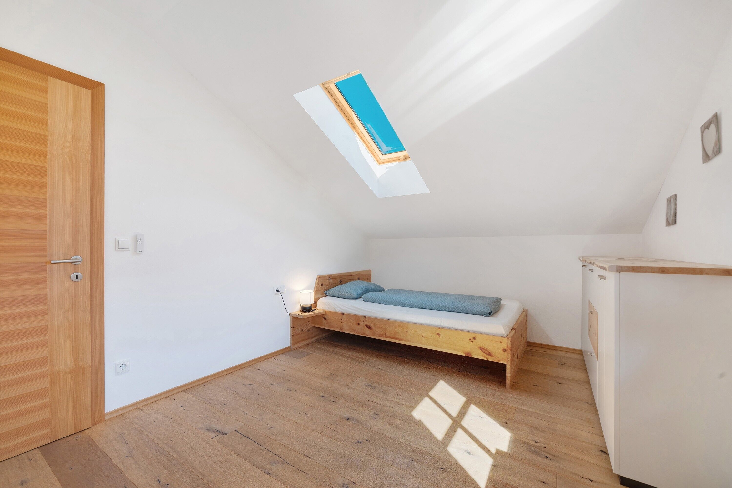 Un piccolo appartamento con una stanza sola, un soffitto spiovente, un letto di legno, una scrivania e un armadio bianco.