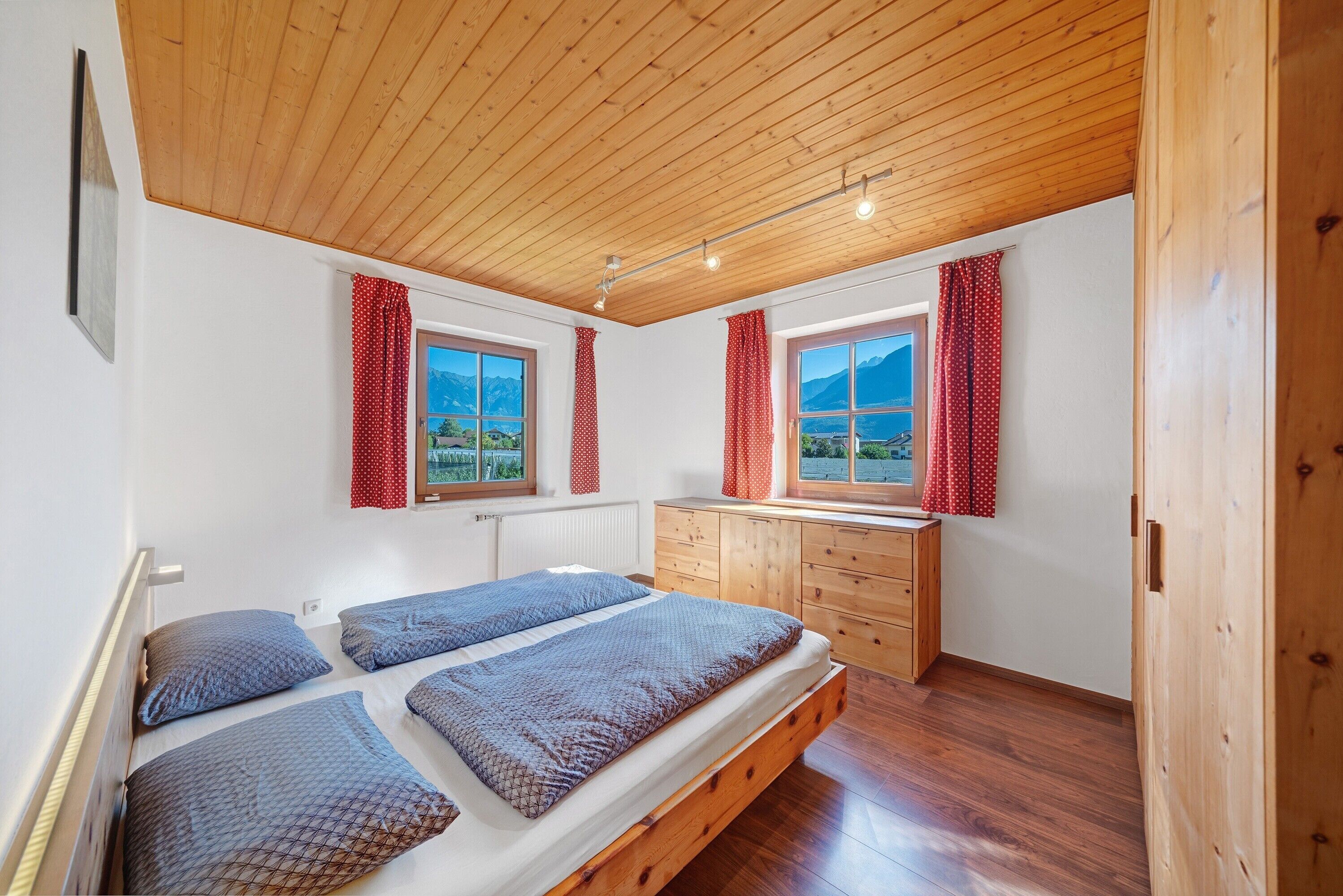 Una camera da letto con soffitto in legno, due finestre con tende, un letto con materasso e cuscini, un comò in legno e vista sulle montagne.
