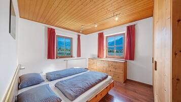 Una camera da letto con soffitto in legno, due finestre con tende, un letto con materasso e cuscini, un comò in legno e vista sulle montagne.