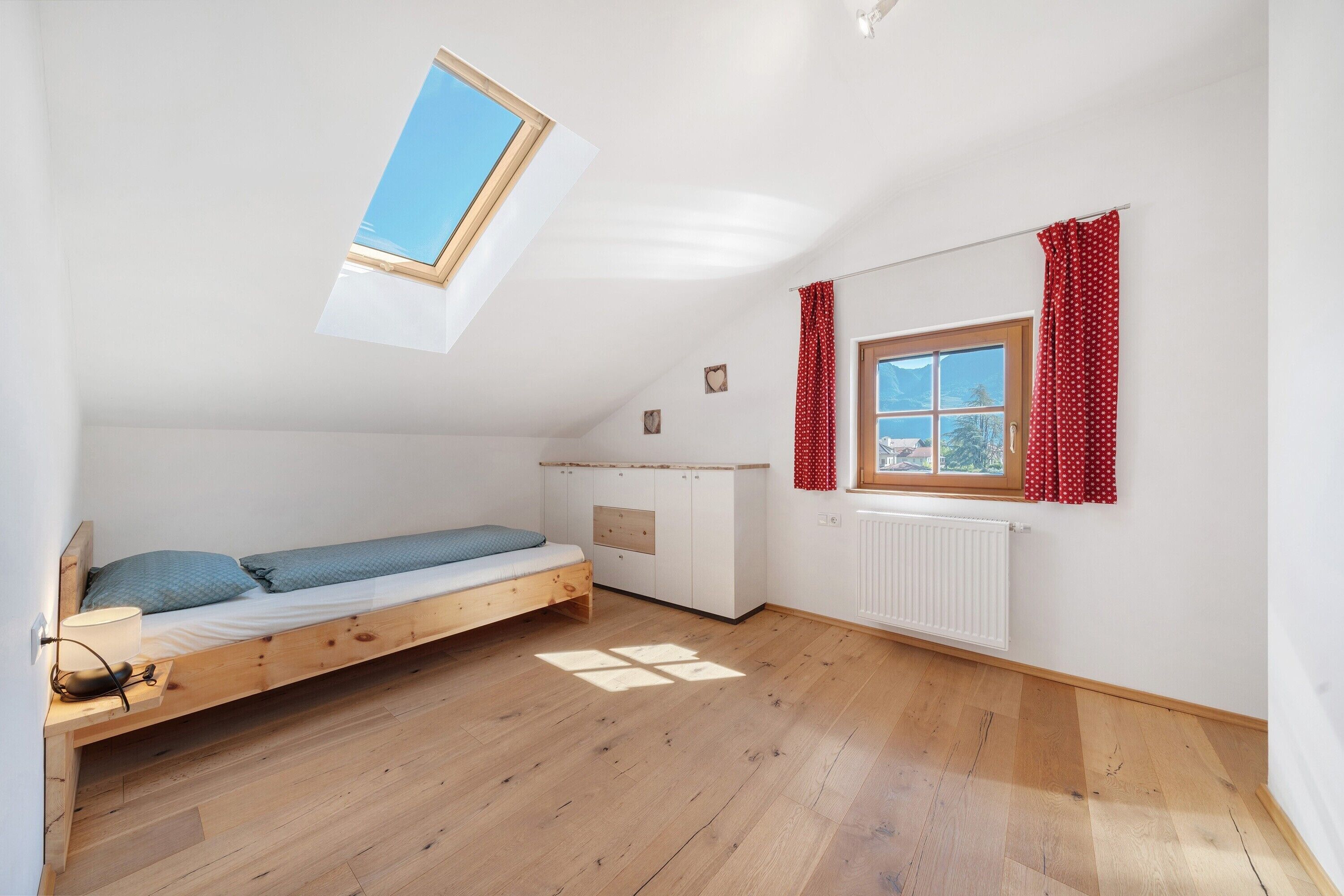 A small, single-room apartment with a sloped ceiling, a bed, a window with curtains, and a radiator.